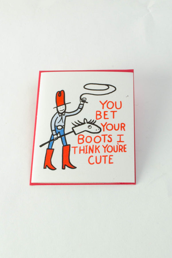 A white greeting card with an illustrated stick-horse rider swinging a lasso. The red text reads "you bet your boots I think you're cute."