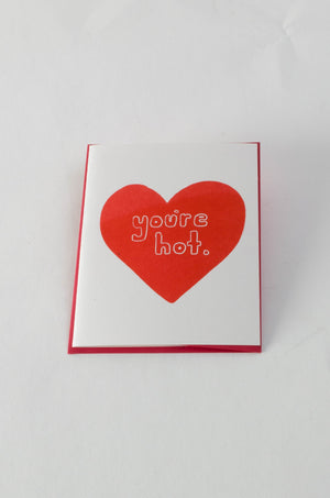 A white greeting card with a large red heart in the center. The text over the heart reads "you're hot."