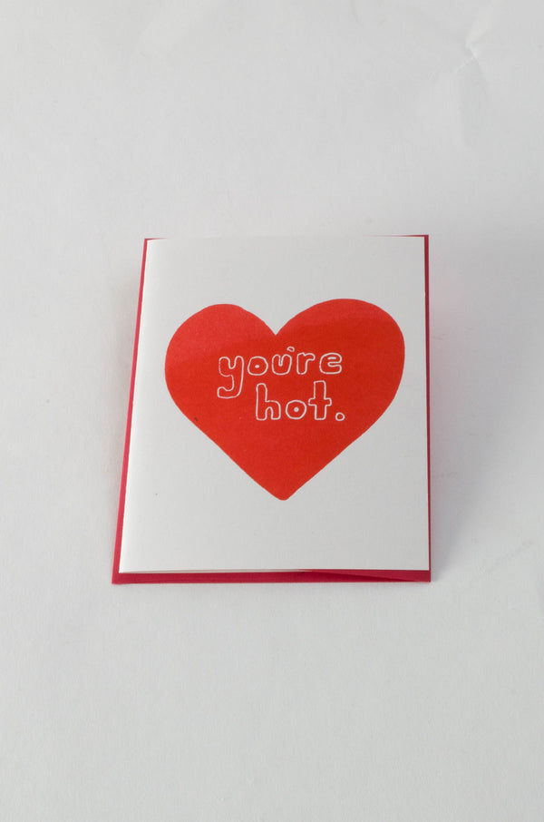A white greeting card with a large red heart in the center. The text over the heart reads "you're hot."