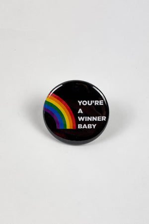 A black pinback button with a rainbow on the left side and white text that reads "you're a winner baby."