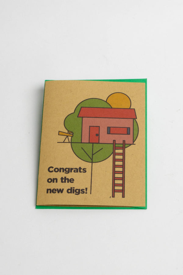 Kraft brown card with green envelope on a white background. The card design features a red tree house with a red ladder and telescope. Below the illustration the card says Congrats on the new digs!