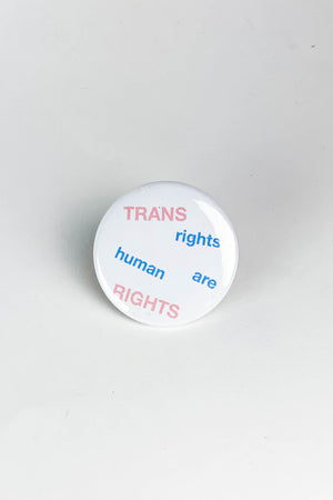 Round pinback button on white background. The button is white with pink and blue text that says Trans rights are human rights. The words are spread out in alternating straight and diagonal patterns.