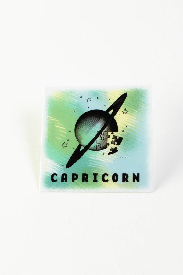 A square sticker with shades of green, blue, and yellow depicting an illustration of the planet Saturn comprised of puzzle pieces. The text reads "Capricorn."