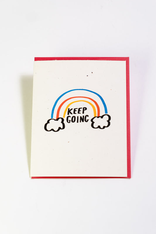 An off-white greeting card with a rainbow emerging from 2 clouds. The black text reads "Keep Going."