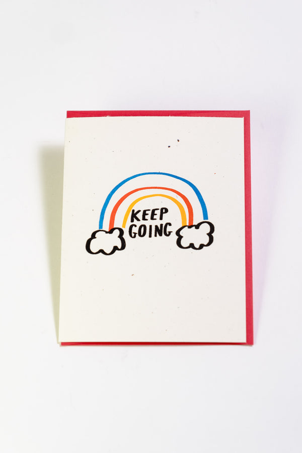 An off-white greeting card with a rainbow emerging from 2 clouds. The black text reads "Keep Going."