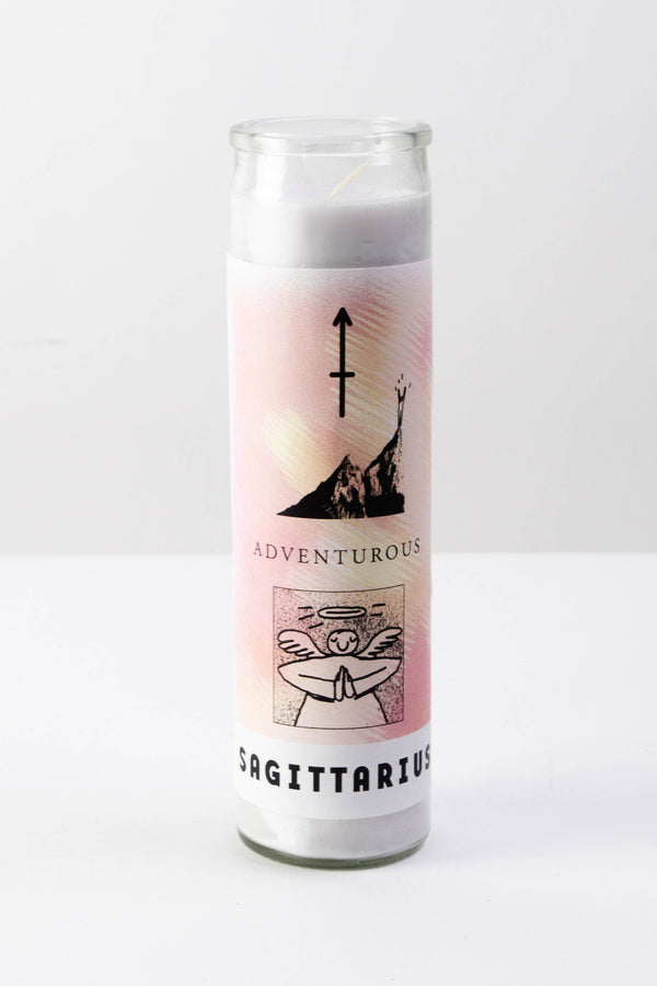 A yellow and pink decal is affixed to a tall glass votive candle. The candle is sitting on a flat white surface. The decal has illustrations symbols attributed to the zodiac sign Sagittarius and features text that reads "Sagittarius," "Adventurous."