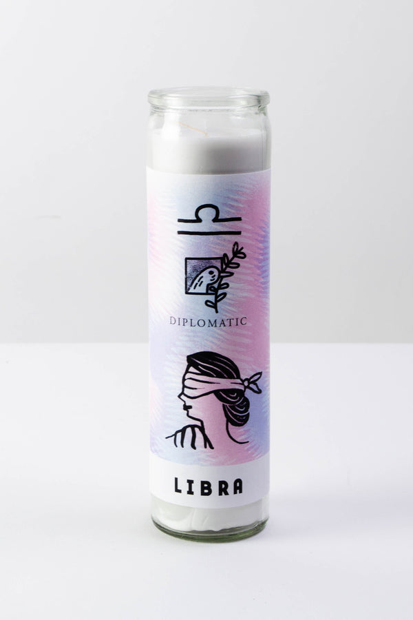 A tall glass votive candle with a pink, blue, and white sticker depicting the astrological symbol for Libra, a dove with an olive branch, the words "Diplomatic" and "Libra" in black text, and a blindfolded woman.