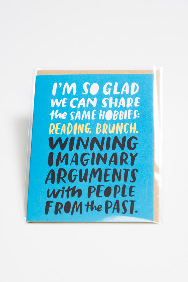 A light blue greeting card with white, yellow, and black text that reads "I'm so glad we can share the same hobbies: reading, brunch, winning imaginary arguments with people from the past."