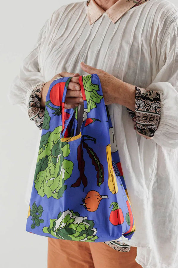 Someone holding a blue reusable shopping tote with illustrations of vegetables in a variety of colors against a white background.