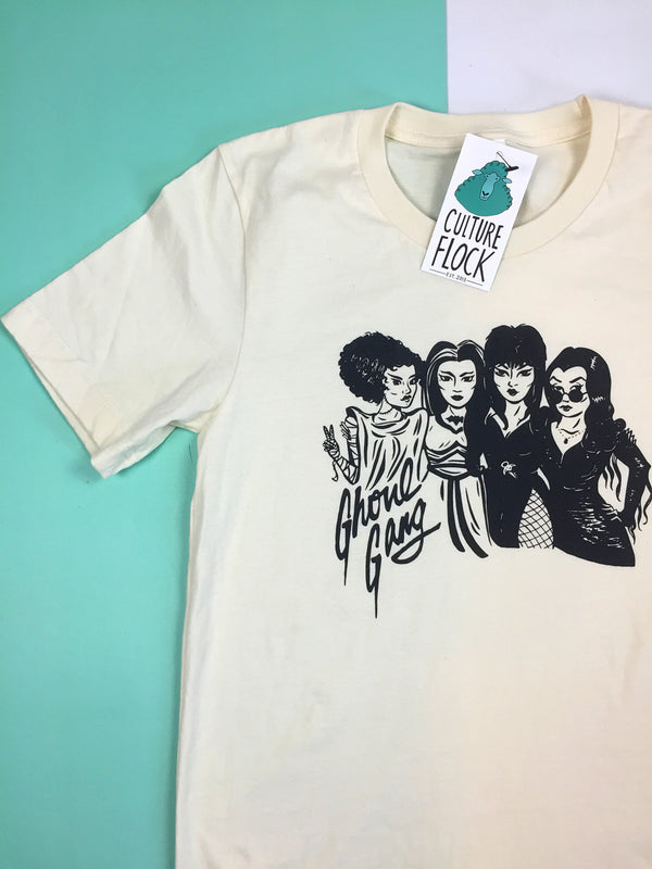 An off-white color t-shirt with black ink featuring illustrations of the Bride of Frankenstein, Lily Munster, Elvira, and Morticia Addams and the words "Ghoul Gang" sits on top of a flat teal and white background.