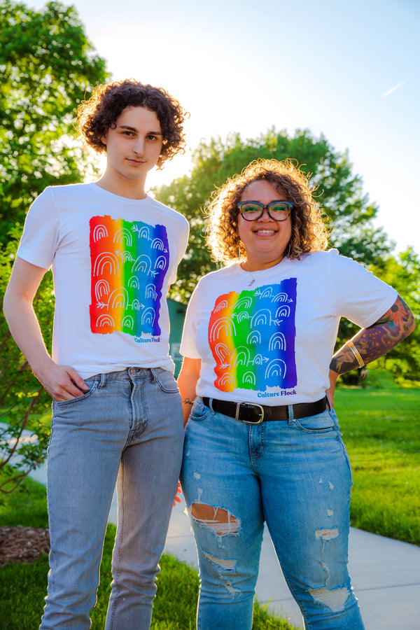 Two people are standing next to a green lawn and sidewalk on a sunny day with large trees behind them. They're both wearing jeans and white t-shirts with a rectangular vertical rainbow and small white illustrated rainbows.