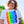 Load image into Gallery viewer, A woman with dark curly hair is standing in the sun in front of green trees. She is wearing jeans, a black belt, and a tucked in white t-shirt with a rectangular vertical rainbow pride flag and white illustrated rainbows overlaying the flag. 
