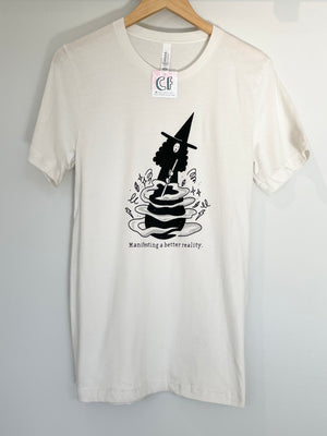 A photograph of an off-white t-shirt on a wooden hanger. The shirt has features an illustration in black ink of a witch stirring the contents of a cauldron. The black text reads "Manifesting a better reality."