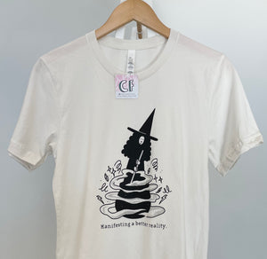 A photograph of an off-white t-shirt on a wooden hanger. The shirt has features an illustration in black ink of a witch stirring the contents of a cauldron. The black text reads "Manifesting a better reality."