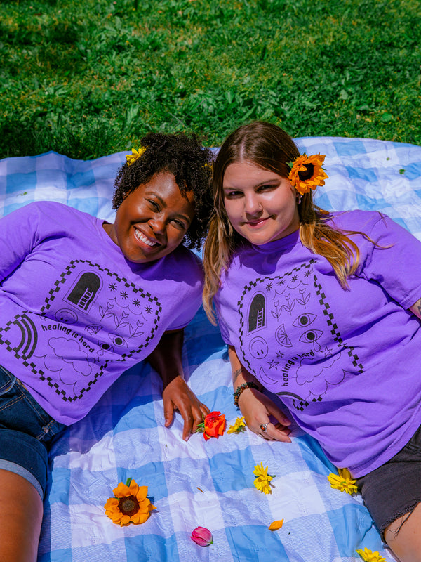 Two women wearing purple "Healing is Hard" t-shirts are laying on a blue checkered picnic blanket in the grass. Flowers are in their hair and some flower petals are scattered on the blanket.