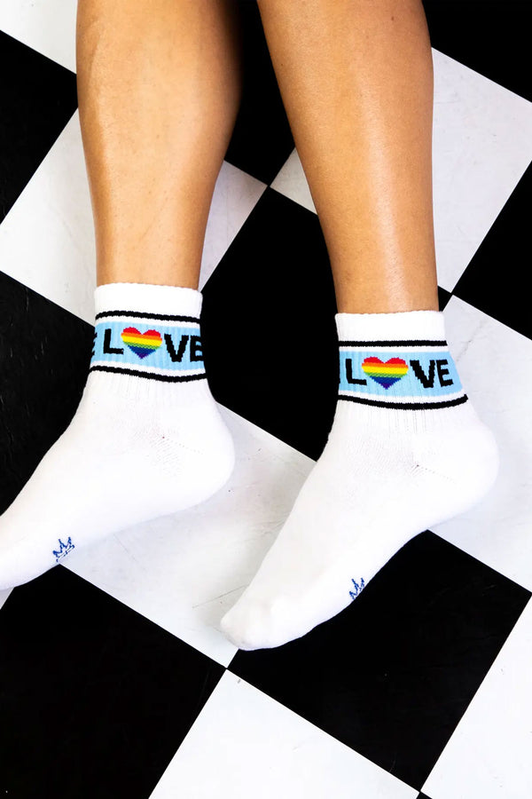A person wearing white ankle crew socks. The socks have a blue stripe with two thin black stripes above and below it. Across the blue stripes in black lettering the socks say LOVE. The O is a heart shape with rainbow colors. Black and white checked background.