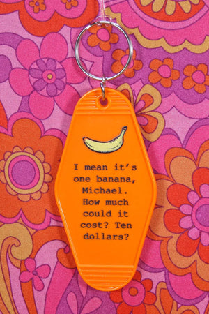 Orange plastic key chain with silver key ring at the top laying on a pink and orange 1960's pattern background. The keychain has a quote from Arrested Development's character Lucille Bluth. It reads "I mean it's one banana Michael. How much could it cost? Ten Dollars?"
