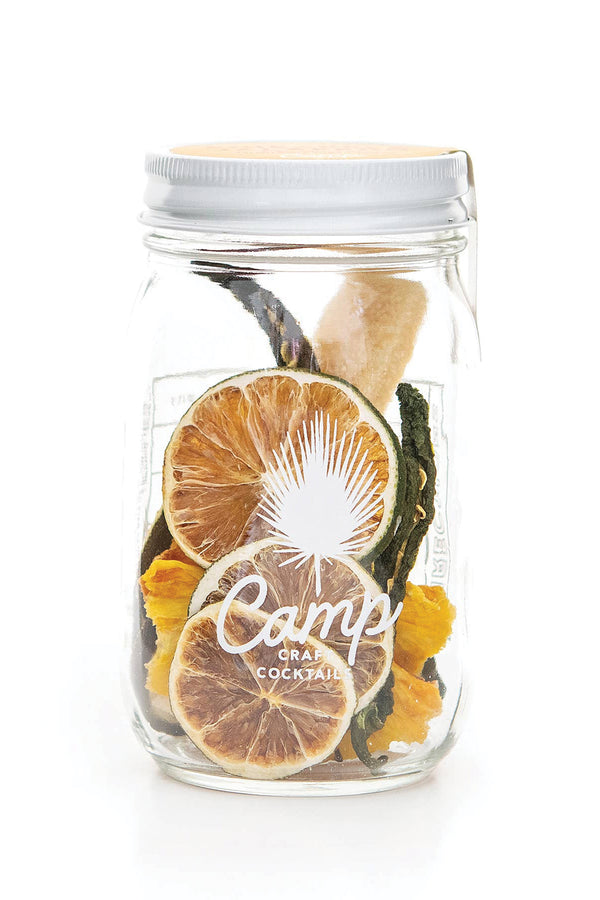 A clear mason jar with a white lid on a white background. The jar is filled with fried fruit and a stick of sugar. The jar features a logo and the text reads "Camp Craft Cocktails."