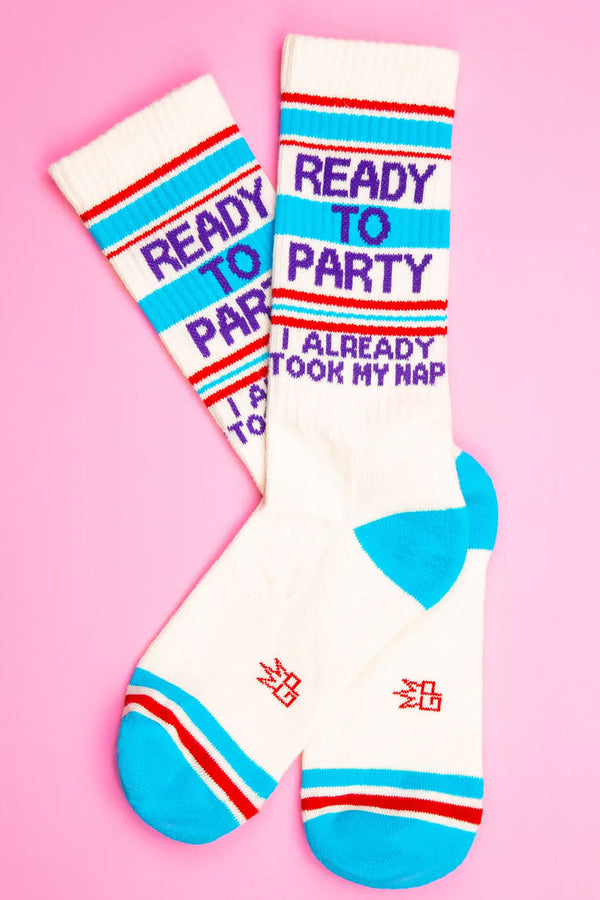 Tall mid-calf crew socks. They have a blue heel and toe. There is a blue and red stripe at the toe and around the stop of the socks. In between the stripes at the top of the socks in purple text, it says "Ready to Party I already took my nap". White background. Pink Background.