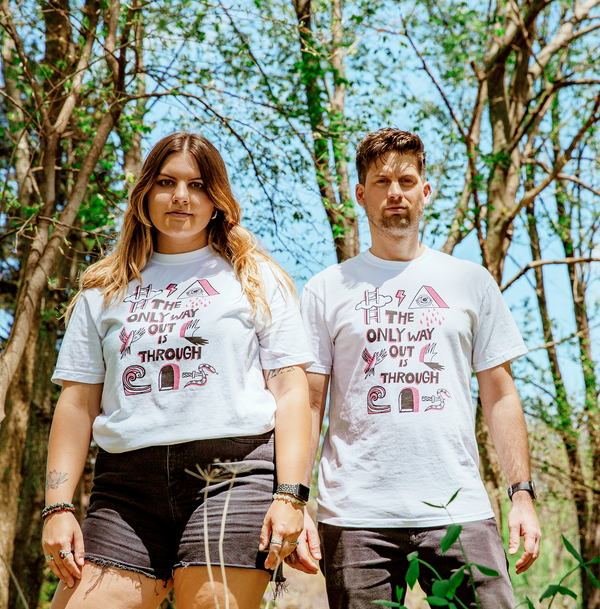 Man and woman outside standing in front of small trees under a blue skies. They are wearing white shirts. The tshirt says "The Only Way Out is Through" in pink and black block letters. Surrounding the text are black and pink illustrations of a ladder going through a cloud, a triangle with a crying eye in the middle, a flying bird, a hand and its shadow, a wave, a snake with a knife piercing it, and a dark arched passageway.