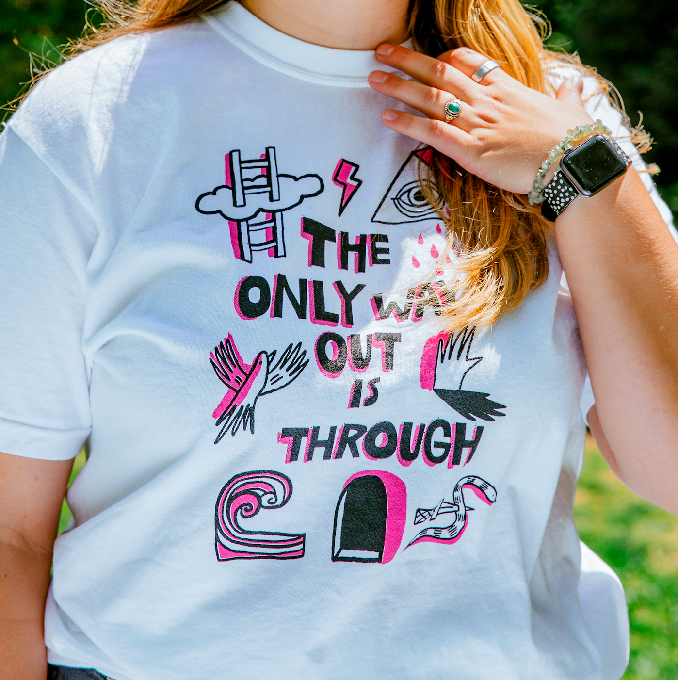 Woman with long hair outside wearing a white tshirt. The tshirt says "The Only Way Out is Through" in pink and black block letters. Surrounding the text are black and pink illustrations of a ladder going through a cloud, a triangle with a crying eye in the middle, a flying bird, a hand and its shadow, a wave, a snake with a knife piercing it, and a dark arched passageway.