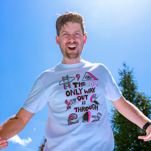 Man outside against a blue sky wearing a white tshirt. The tshirt says "The Only Way Out is Through" in pink and black block letters. Surrounding the text are black and pink illustrations of a ladder going through a cloud, a triangle with a crying eye in the middle, a flying bird, a hand and its shadow, a wave, a snake with a knife piercing it, and a dark arched passageway.