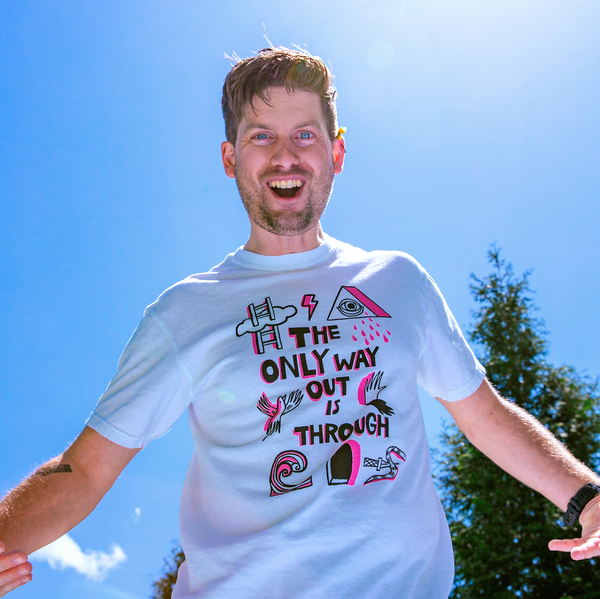 Man outside against a blue sky wearing a white tshirt. The tshirt says "The Only Way Out is Through" in pink and black block letters. Surrounding the text are black and pink illustrations of a ladder going through a cloud, a triangle with a crying eye in the middle, a flying bird, a hand and its shadow, a wave, a snake with a knife piercing it, and a dark arched passageway.