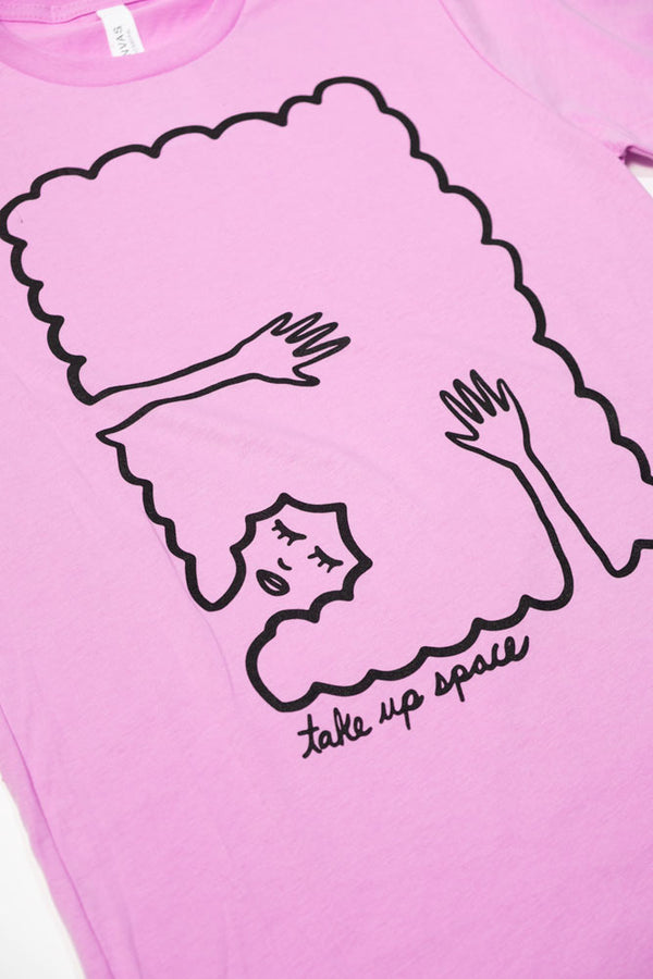 Lilac tshirt on a white background. The Tshirt design is black line work of a person laying down with their hands abover their head laying on their hair. The hair creates a rectangle border. Under the illustration the shirt says Take Up Space.