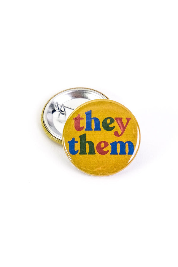 Yellow button laying on top of a turned over pinback button on a white background. The button says "They Them" in alternating letter colors of blue red and green.