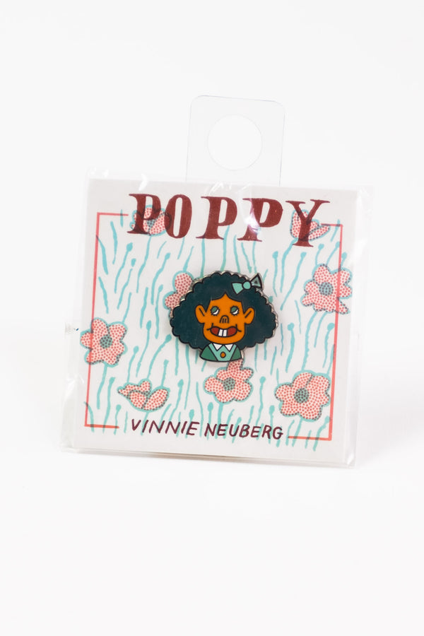 A die-cut enamel pin of a girl with big hair decorated with a green bow-tie, and cartoonish features. The pin is affixed to a cardstock background that has flowers and reads "Poppy" by artist "Vinnie Neuberg."