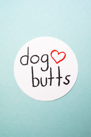 A round white sticker that reads "dog butts" in black with an outline of a red heart. The background is pale blue.