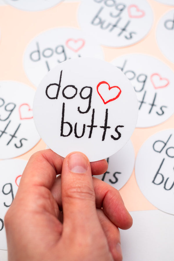 A hand holding a "dog butts" sticker in the foreground of multiple stickers in the background.