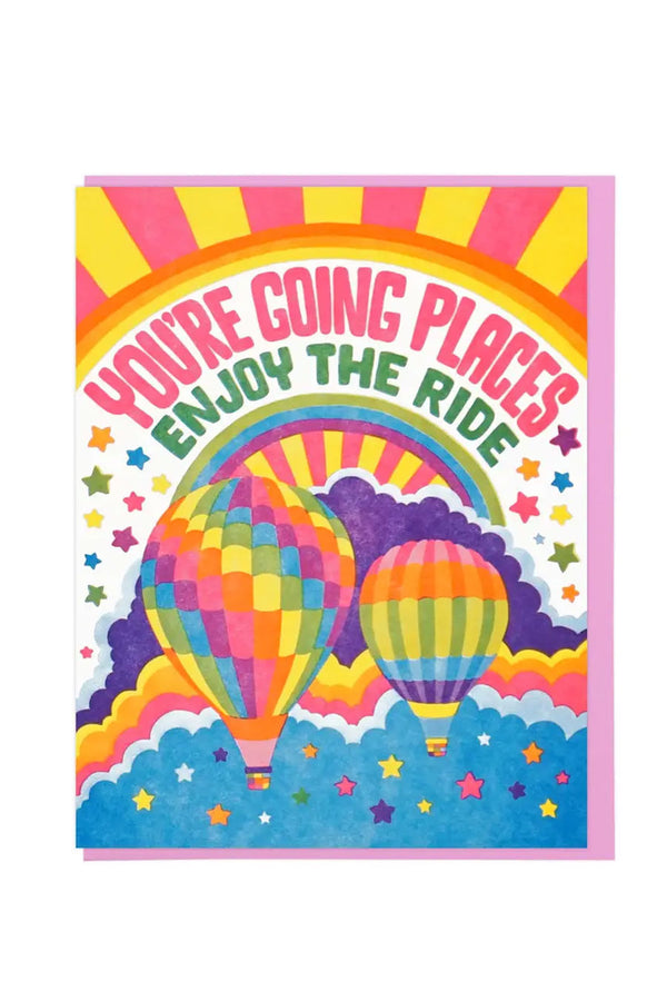 Colorful card with pink and yellow sun burst. Under that the card says "You're Going Places, Enjoy the Ride" in pink and green text. Under the text is two rainbow color hot air balloons hovering above blue clouds. Pink, orange, yellow, green, and blue stars surround the balloon. White background.