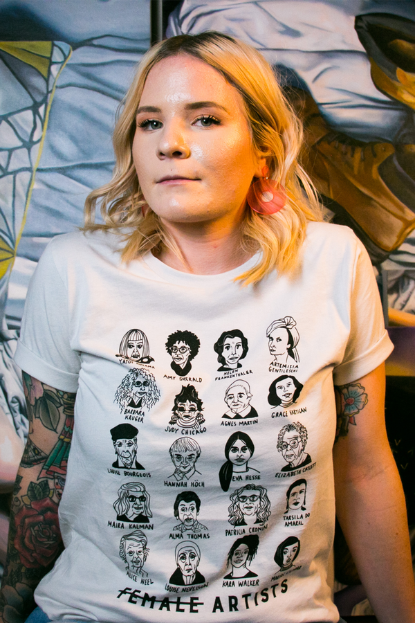 A blonde woman wearing a white tee shirt with illustrated depictions of historically significant female artists. 