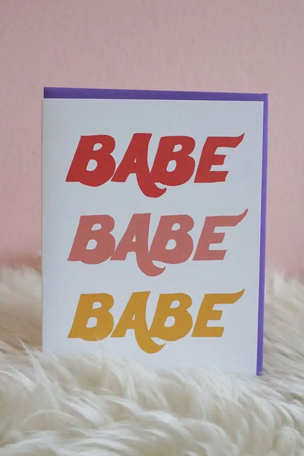 A white greeting card with the words "Babe Babe Babe" written on the front in red, pale pink, and gold letters. Accompanying envelope is purple. The card is sitting in front of light pink background and sitting on white furry rug. 