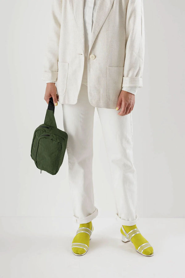 Person wearing white jacket, white shirt, and white pants holding dark green fanny pack with black straps in front of a white background.