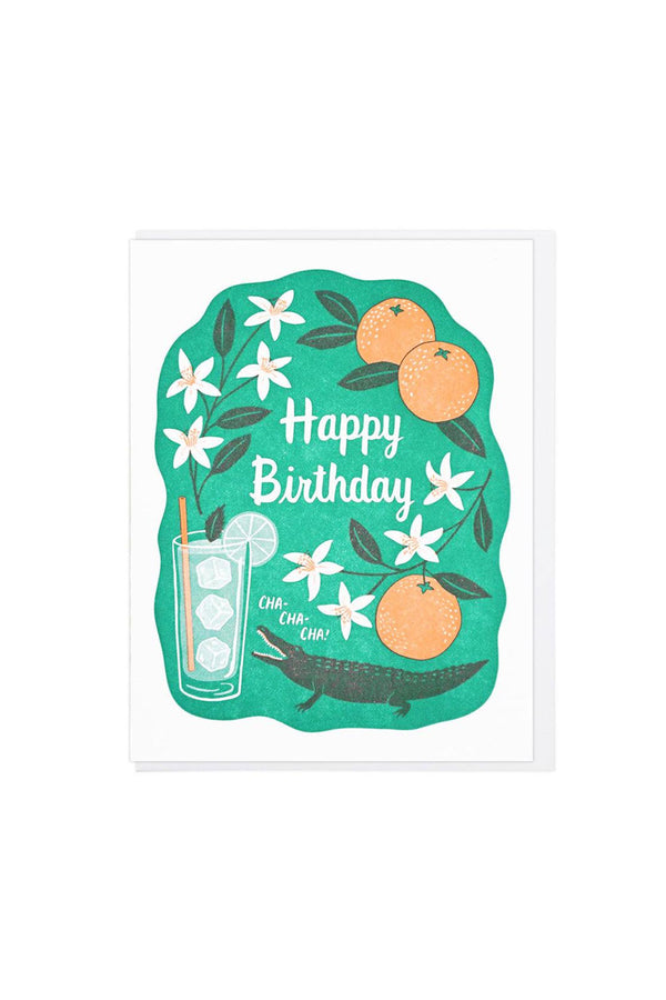 White card with white envelope on a white background. The card features a green shape that has a crocodile next to a glass of ice water with a leaf and a lime slice. Above the crocodile surrounding white text are oranges and orange blossoms. The card says Happy Birthday.