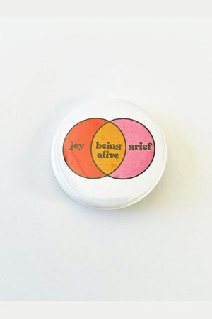 A white pinback button with a venn diagram in red, yellow, and pink. The red left circle is labeled joy, the middle circle is yellow and labeled "being alive", and the right pink circle is labeled "grief."