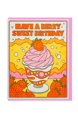 White greeting card with a pink envelope on a white envelope. The card features pink ice cream with a strawberry on top in a white ice cream cup. The cup has orange swirls with pink and yellow shapes throughout. The card has a thin pink border with strawberry clusters on the bottom left and right. Behind the ice cream are yellow and pink cloud shapes. The card says Have a Berry Sweet Birthday in red above the illustration. 