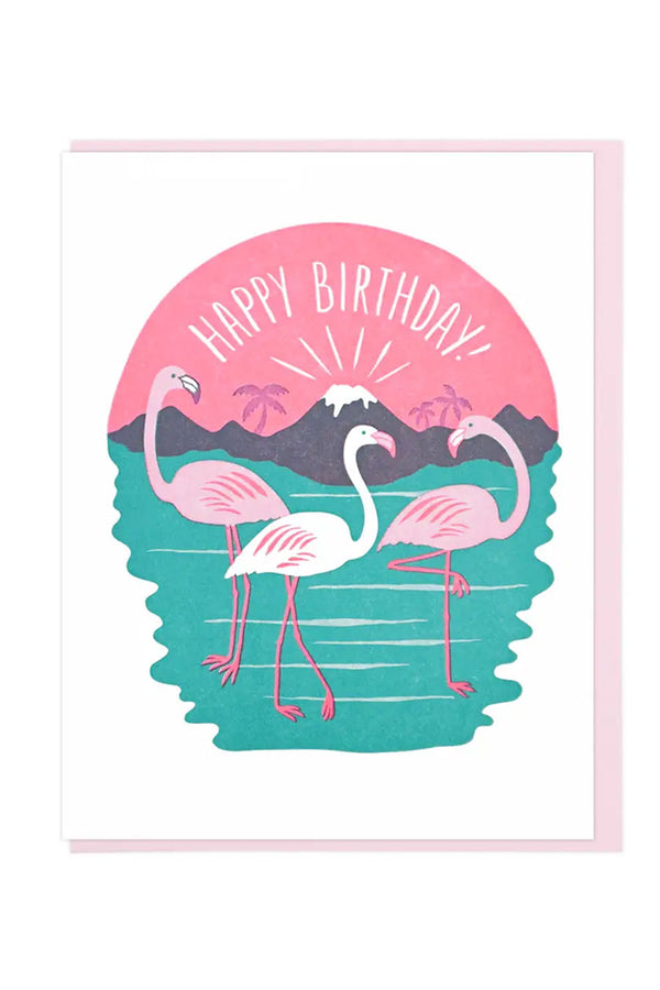White card with a light pink envelope on a white background. The card features three flamingos standing in water in front of a volcano with a pink sky. The card says Happy Birthday in white text against the pink sky.