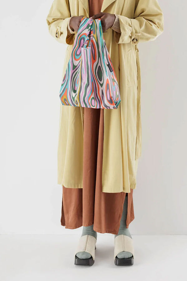 A person wearing a long khaki trench coat and rust color dress against a white backdrop. They are holding a small reusable bag with multicolor ovals and stripes mimicking woodgrain pattern. 