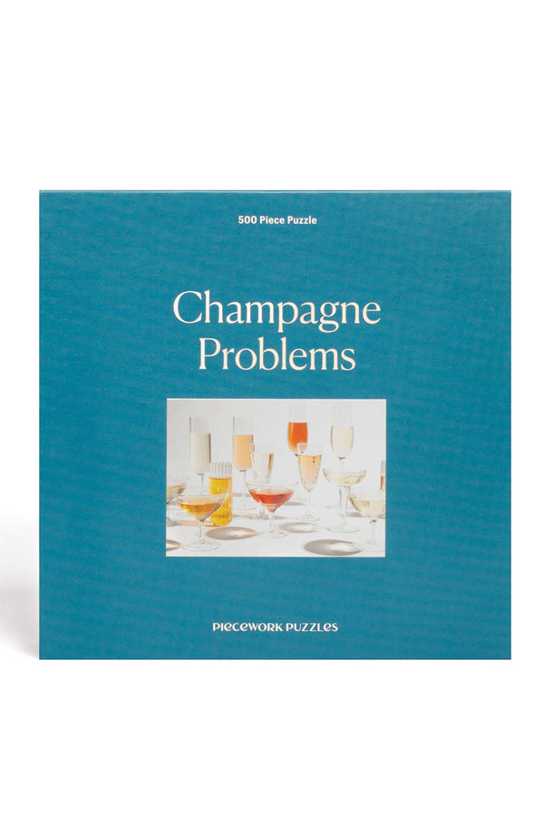 A teal blue jigsaw puzzle box. The puzzle is called "Champagne Problems" and has a photo of several glasses of champagne arranged on a table.