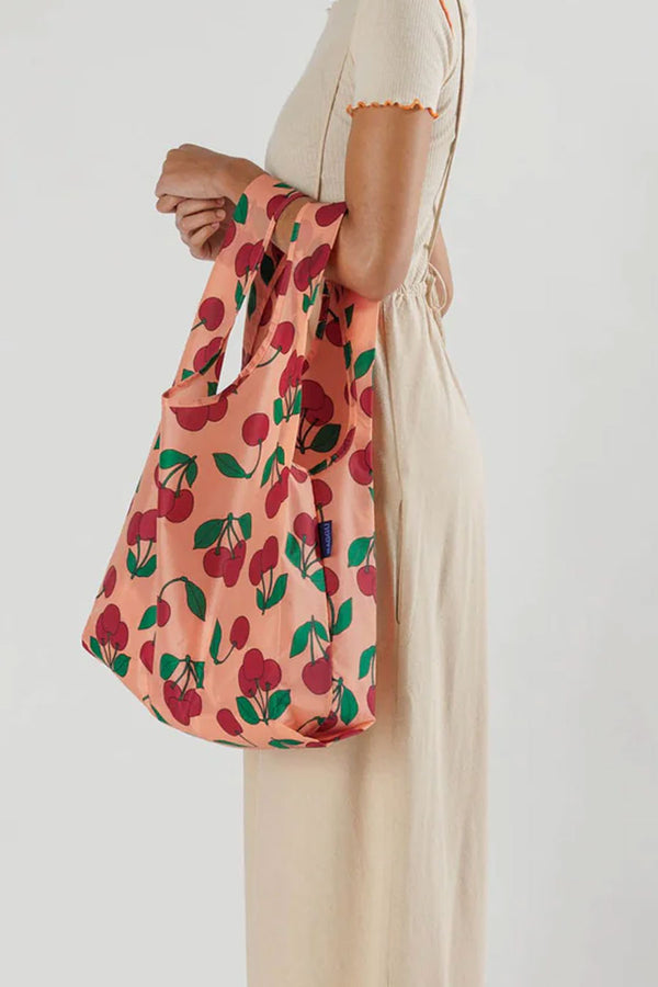 Person against a white background holding a large reusable tote back on their arm. The design of the tote bag is Cherries with stems all over it. 