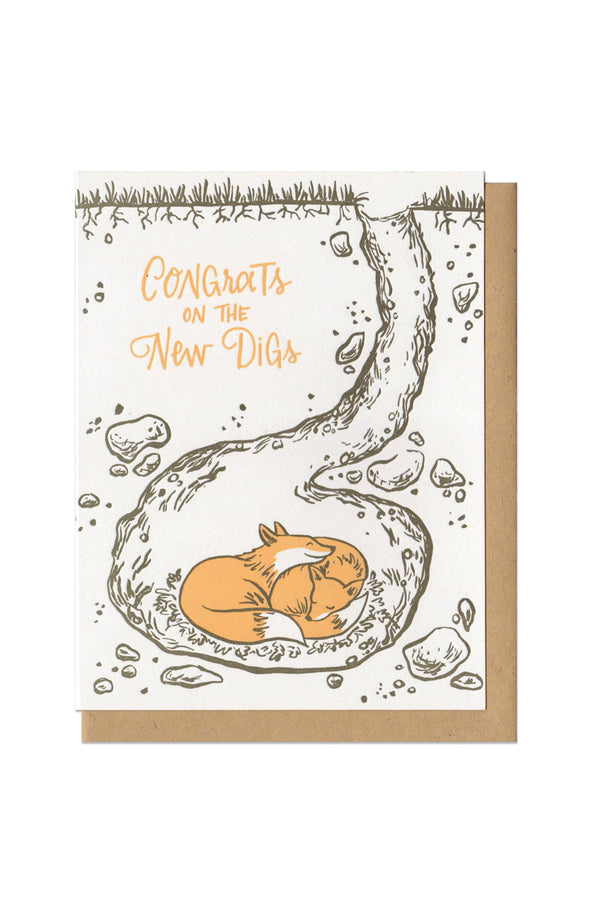 White card with kraft brown envelope on white background. The card illustrates two foxes sleeping in an underground den. The foxes are orange, while the rest of the illustration is a gray brown color. The text in orange says Congrats on the New Digs. 