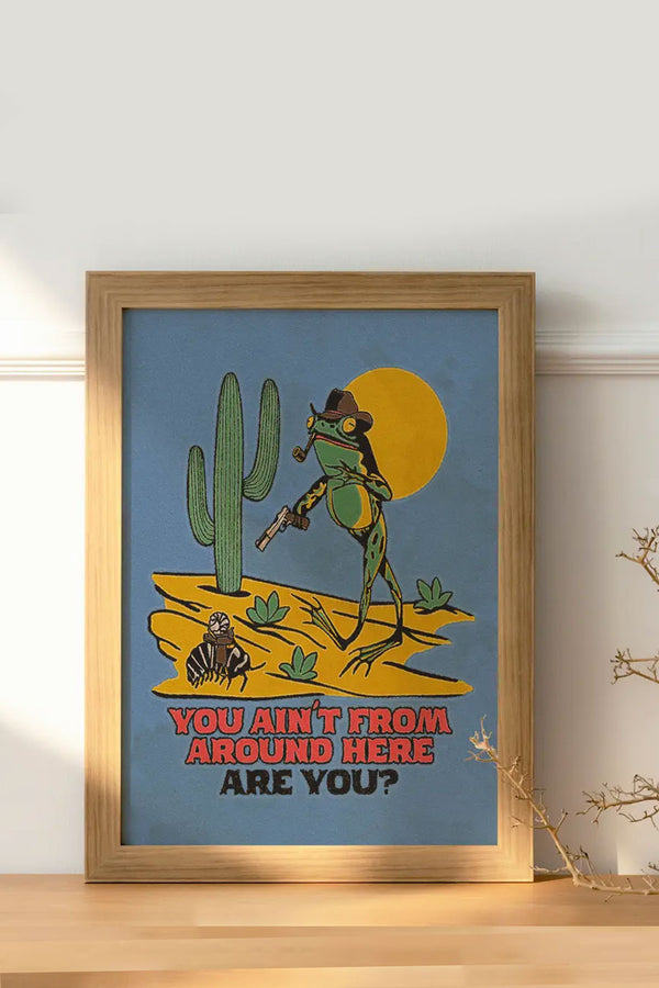 Blue poster in wooded frame on a wooden table leaned up against a white wall. The artwork is a Green toad wearing a cowboy hat standing next to a cactus. The toad has a pipe in his mouth and is pointing a pistol at a desert bug sitting in a saddle on top of a larger desert bug. Below the illustration the poster says "You aint from around here are you?" in red and black western text.