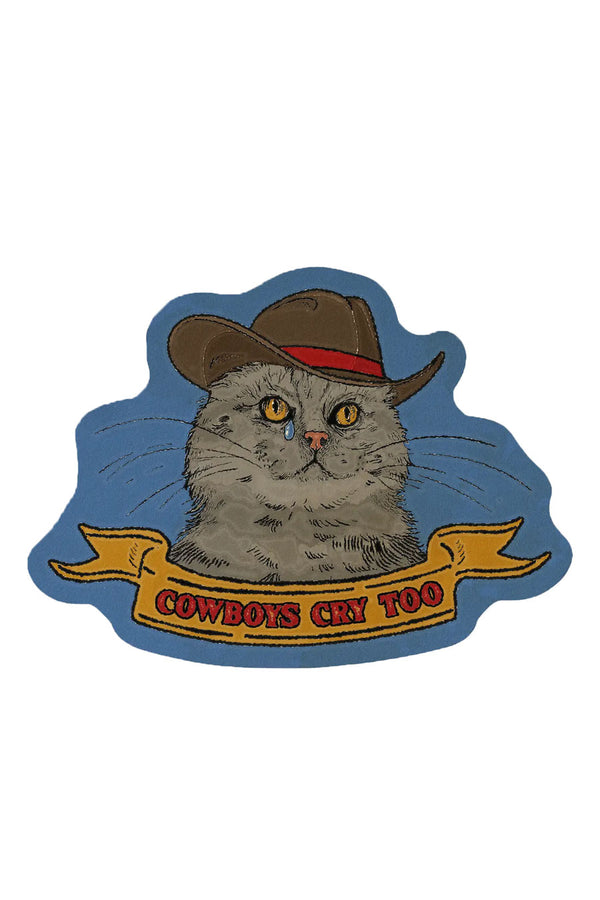 A die-cut sticker of a gray cat wearing a cowboy hat, shedding a tear. The background is a dusty blue. The yellow banner below him says "Cowboys cry too" in red text. 