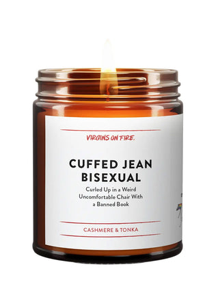 Amber glass candle jar with white label. The label says Cuffed Jean Bisexual, Culled up in a weird uncomfortable chair with a banned book. Scent is Cashmere and Tonka. Brand is Virgins on Fire.