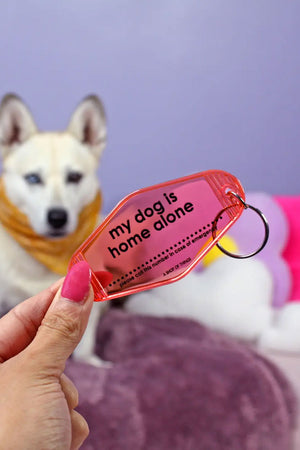 Person holding a translucent Pink motel style keychain. The keychain says My dog is home alone, please call this number in case of emergency. There is a space in between the sentences for you to write a phone number. Purple wall with a white dog in the background.