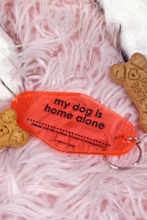 a translucent Pink motel style keychain. The keychain says My dog is home alone, please call this number in case of emergency. There is a space in between the sentences for you to write a phone number.  The keychain is laying on a fuzzy pink blanket surrounded buy dog treats and dog paws.
