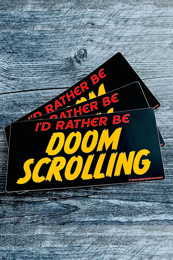 A stack of three black rectangular bumper stickers that read "I'd rather be doom scrolling" in bold yellow and red text on a gray wooden background.